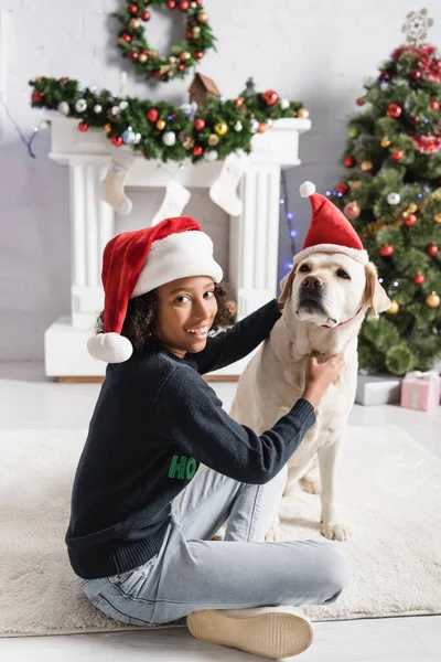 Smiling african american girl cuddling labrador in santa hat near christmas tree on blurred background — Stock Photo