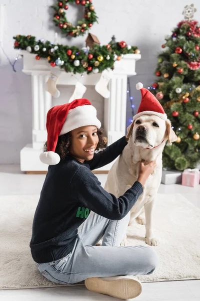 Souriant afro-américaine fille câlinant labrador dans santa chapeau près de l'arbre de Noël sur fond flou — Photo de stock