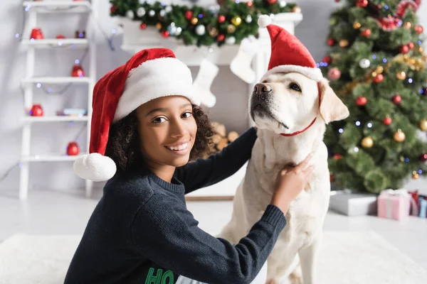 Glücklich afrikanisch-amerikanische Mädchen kuschelt Labrador in Weihnachtsmütze in der Nähe von Weihnachtsbaum auf verschwommenem Hintergrund — Stockfoto