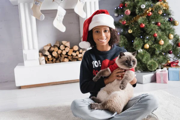 Счастливая африканская американская девушка в шляпе Санты держа кота, сидя возле рождественской елки и камина — стоковое фото