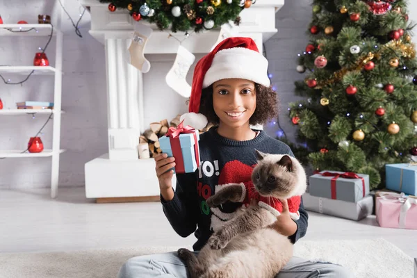 Alegre menina afro-americana em santa chapéu segurando caixas de presente enquanto sentado com gato perto da árvore de natal — Fotografia de Stock