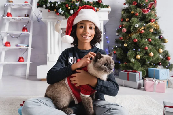 Счастливая африканская американская девушка смотрит в камеру, сидя возле рождественской елки и гладя кошку на размытом фоне — стоковое фото