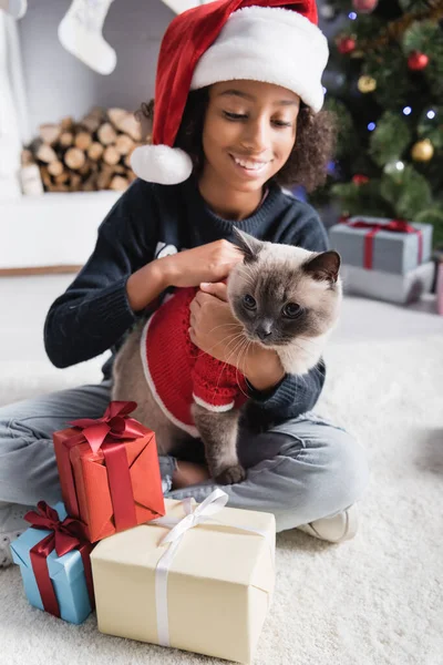 Allegra ragazza afroamericana in cappello di Babbo Natale coccole gatto soffice mentre seduto vicino regali di Natale su sfondo sfocato — Foto stock