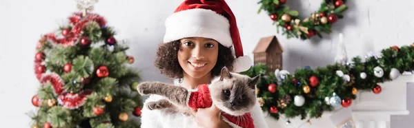 Lächelnde afrikanische Amerikanerin, die in die Kamera schaut, während sie Katze auf verschwommenem Hintergrund mit Weihnachtsdekoration hält, Banner — Stockfoto