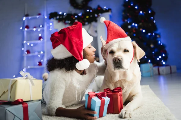 Lächelnde Afroamerikanerin streichelt Labrador-Hund, während sie auf dem Boden in der Nähe von Geschenkboxen auf verschwommenem Hintergrund liegt — Stockfoto