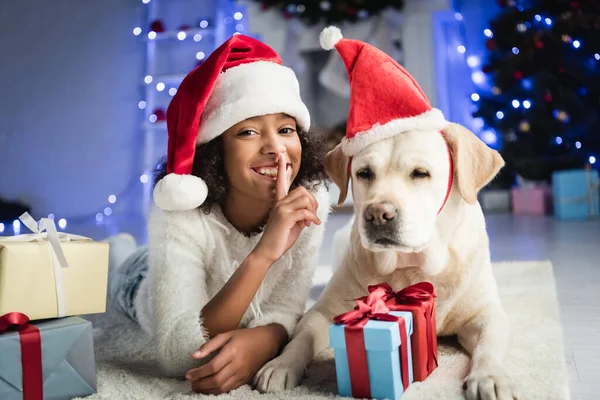 Africano americano ragazza mostrando silenzio gesto mentre sdraiato su pavimento vicino labrador e regalo scatole su sfondo sfocato — Foto stock