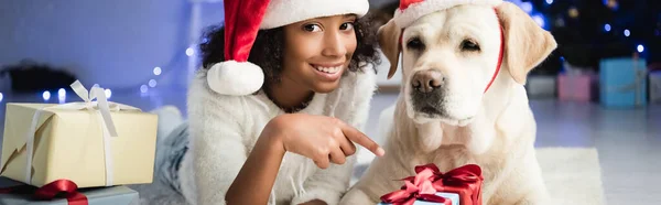 Happy african american girl pointing with finger at gift box while lying on floor near labrador dog on blurred background, banner — Stock Photo