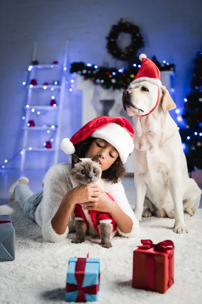 Africana chica americana acurrucándose gato mientras está acostado en el suelo cerca de labrador perro y regalos de Navidad sobre fondo borroso - foto de stock