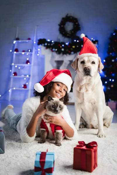 Cheerful african american girl cuddling fluffy cat while lying on floor near labrador dog on blurred background — Stock Photo