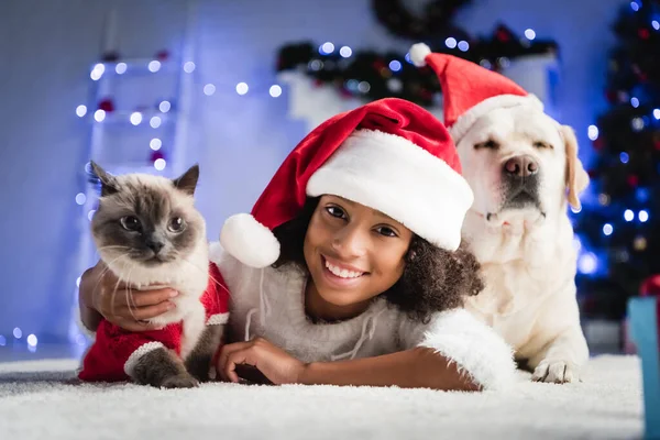 Happy african american girl in santa hat lying on floor near labrador and cat on blurred background — Stock Photo