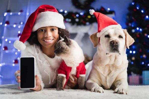 Fille souriante montrant smartphone avec écran vide et couché sur le sol près du chat et récupérateur sur fond flou — Photo de stock