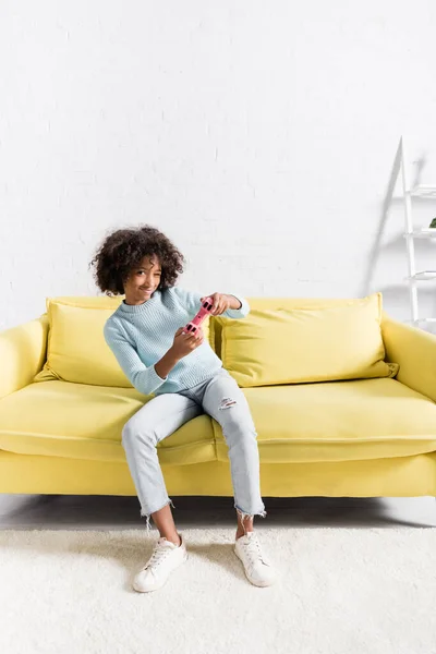 KIEW, UKRAINE - 02. OKTOBER 2020: Fröhliches afrikanisch-amerikanisches Mädchen spielt mit rosa Joystick auf gelbem Sofa zu Hause — Stockfoto