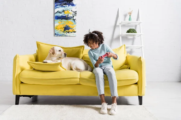 KYIV, UKRAINE - OCTOBER 02, 2020: Preteen girl playing with joystick and sitting near labrador on sofa at home — Stock Photo