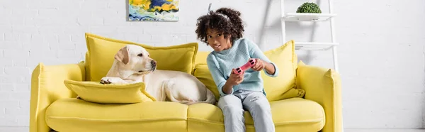 KIEW, UKRAINE - 02. OKTOBER 2020: Konzentriertes Mädchen, das am Steuerknüppel spielt, während es neben Labrador auf dem heimischen Sofa sitzt, Banner — Stockfoto