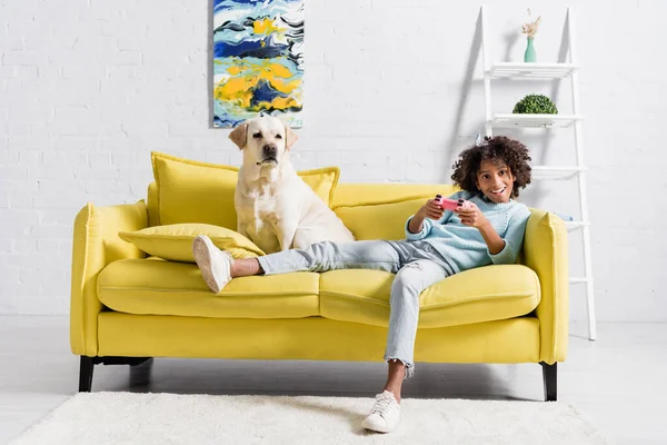 KYIV, UKRAINE - OCTOBER 02, 2020: Labrador sitting near cheerful girl playing with joystick on yellow sofa at home - foto de stock