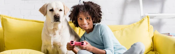 KYIV, UCRANIA - 02 DE OCTUBRE DE 2020: niña afroamericana sonriente sentada cerca de labrador, mientras juega con el joystick en casa, pancarta - foto de stock