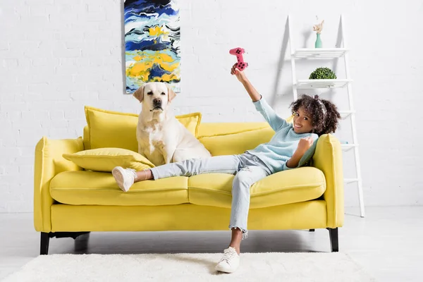 KYIV, UKRAINE - OCTOBER 02, 2020: cheerful curly girl with pink joystick lying on sofa near retriever at home — Foto stock