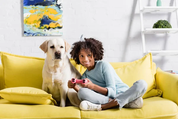 QUIIV, UCRÂNIA - OUTUBRO 02, 2020: menina afro-americana sentada perto do labrador, enquanto brincava com joystick em casa — Fotografia de Stock