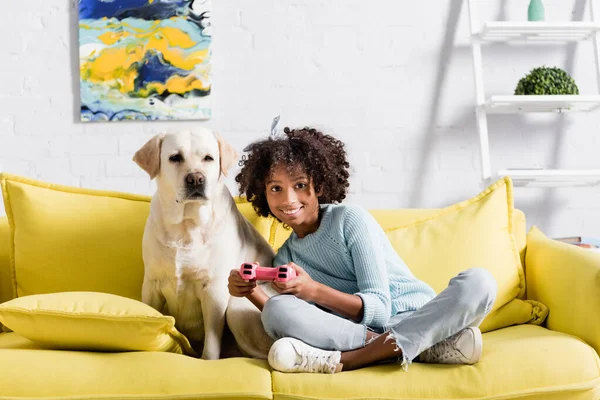 KIEW, UKRAINE - 02. OKTOBER 2020: Afrikanerin sitzt neben Labrador und spielt zu Hause mit Joystick — Stockfoto