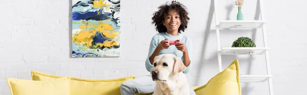 KYIV, UKRAINE - 02 OCTOBRE 2020 : fille heureuse assise derrière le labrador sur le canapé, tout en jouant avec joystick à la maison, bannière — Photo de stock