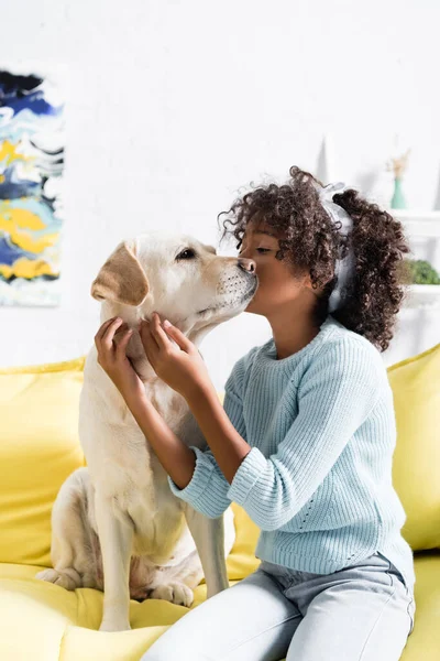 African american girl with headband, kissing labrador muzzle, while sitting near on yellow sofa at home — Stock Photo