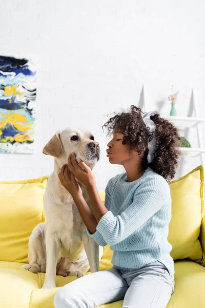 Afro-américaine fille avec des lèvres boudantes, toucher muselière labrador, tout en étant assis près sur canapé jaune à la maison — Photo de stock