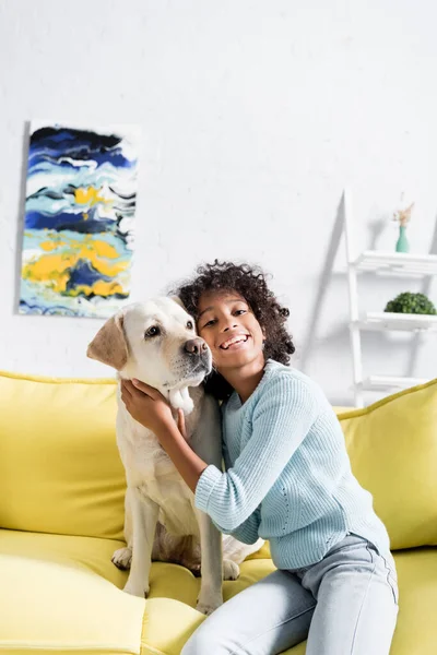 Curly african american girl embracing retriever, while sitting on yellow sofa at home, on blurred background — Stock Photo