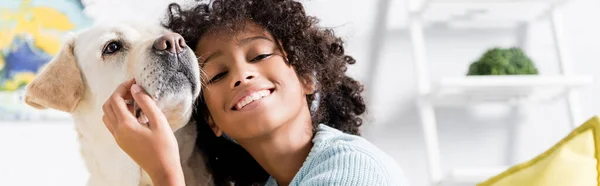 Happy african american girl leaning on retriever at home on blurred background, banner — Stock Photo