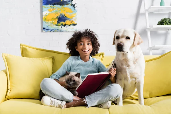 Feliz chica afroamericana rizada con libro abierto, abrazando al gato, mientras está sentada cerca de retriever en el sofá en casa - foto de stock