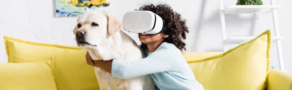 Afroamericano ragazza in auricolare vr abbracciare cane, mentre seduto sul divano giallo a casa, banner — Foto stock