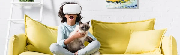 Africano menina americana com boca aberta em vr fone de ouvido abraçando gato, enquanto sentado no sofá em casa, banner — Fotografia de Stock