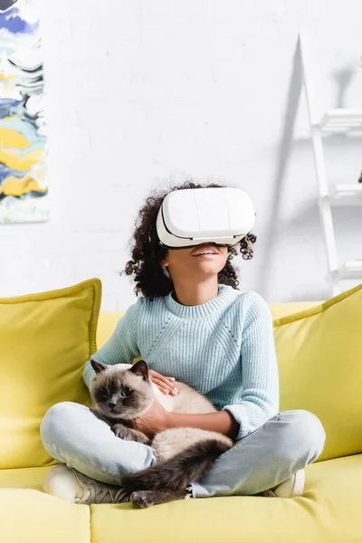 Smiling african american girl with crossed legs wearing vr headset, stroking cat and sitting on sofa on blurred background — Stock Photo