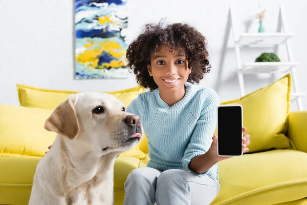 Африканская американка показывает смартфон с пустым экраном и сидит рядом с лабрадором на диване на размытом фоне — стоковое фото