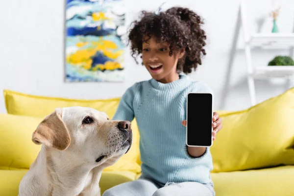 Retriever sitzt neben lächelndem afrikanisch-amerikanischen Mädchen und zeigt Smartphone mit leerem Bildschirm auf verschwommenem Hintergrund — Stockfoto