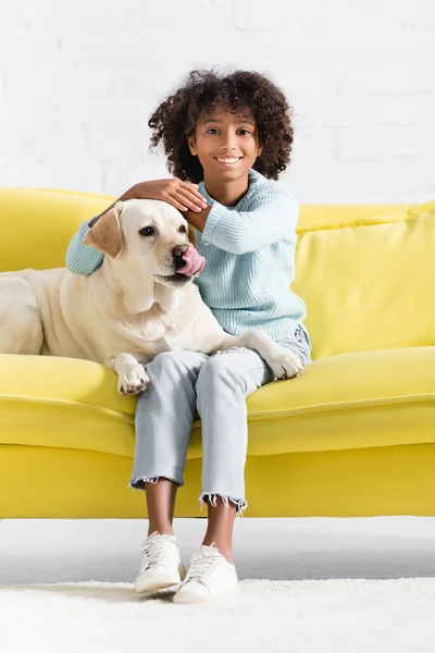 Lächelnde Afroamerikanerin blickt in die Kamera, während sie neben Labrador sitzt, der sich zu Hause auf dem Sofa leckt — Stockfoto