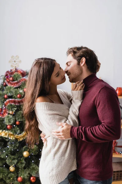 Щаслива пара в светрах цілується біля прикрашеної ялинки на розмитому фоні — стокове фото