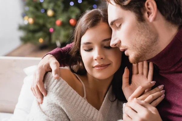 Casal satisfeito em camisolas aconchegantes abraçando perto da árvore de natal decorada no fundo borrado — Fotografia de Stock