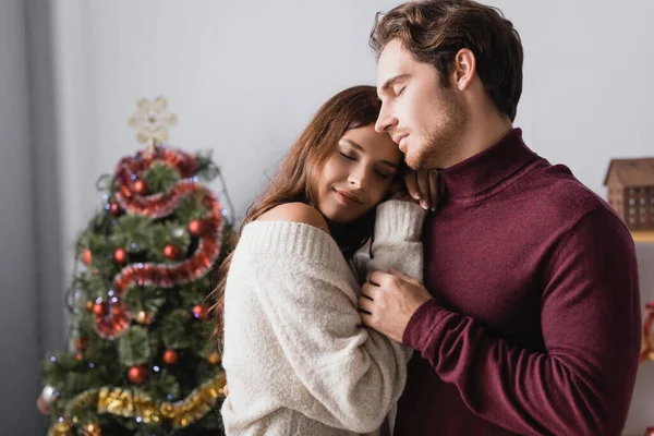 Coppia in maglioni caldi abbracciando vicino decorato albero di Natale su sfondo sfocato — Foto stock