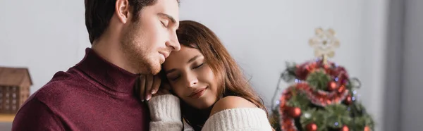 Pareja en suéteres calientes abrazándose cerca del árbol de Navidad decorado sobre un fondo borroso, pancarta - foto de stock