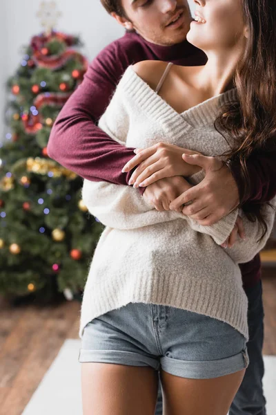 Vue recadrée du couple heureux dans des pulls chauds étreignant près de l'arbre de Noël décoré sur fond flou — Photo de stock