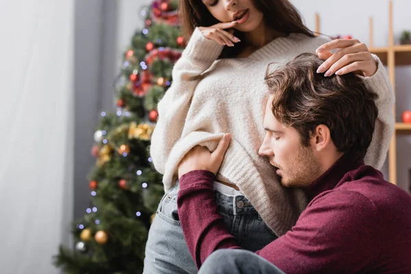 Passionate man undressing young woman in sweater near christmas tree on blurred background — Stock Photo