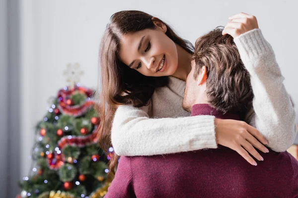 Homme passionné embrasser jeune femme heureuse en pull près de l'arbre de Noël sur fond flou — Photo de stock