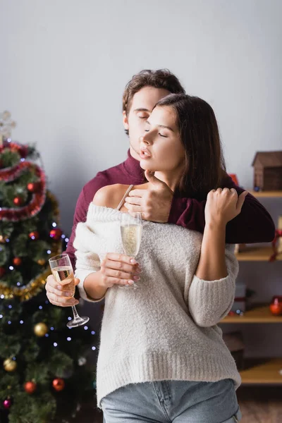 Passionate man embracing woman while holding glasses with champagne near christmas tree on blurred background — Stock Photo