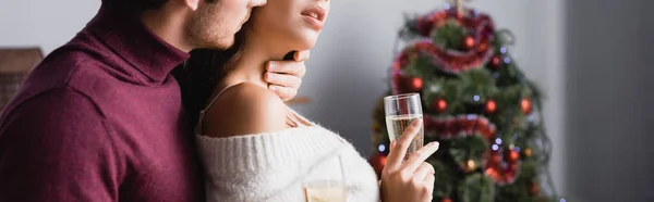 Corte vista de homem apaixonado beijando mulher e segurando óculos com champanhe perto da árvore de natal no fundo borrado, banner — Fotografia de Stock