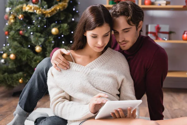 Paar in Pullovern mit digitalem Tablet in der Nähe des Weihnachtsbaums auf verschwommenem Hintergrund — Stockfoto