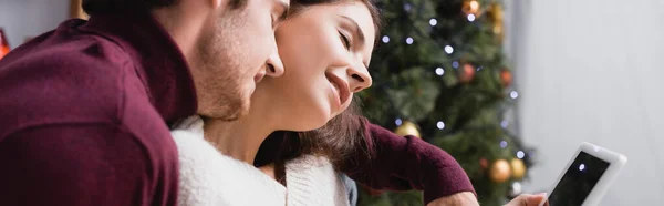 Frau im Pullover hält digitales Tablet mit leerem Bildschirm neben Freund und Weihnachtsbaum auf verschwommenem Hintergrund, Banner — Stockfoto