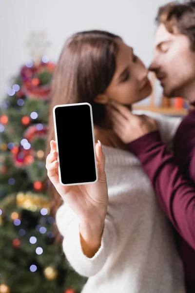 Mujer sosteniendo teléfono inteligente con pantalla en blanco cerca de novio y árbol de Navidad decorado sobre fondo borroso - foto de stock