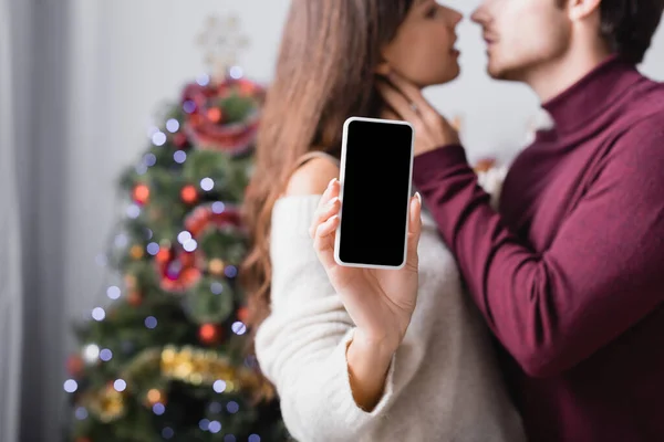 Женщина держит смартфон с пустым экраном рядом с парнем и елкой на размытом фоне — стоковое фото