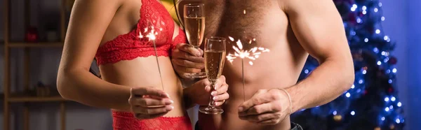 Vista recortada de pareja sexy sosteniendo copas con champán y bengalas cerca de la iluminación borrosa, pancarta - foto de stock