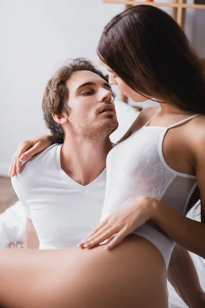 Sexy woman in bodysuit embracing boyfriend with closed eyes in bedroom — Stock Photo